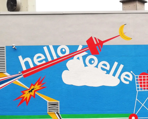 Ausschnitt der mehrfarbigen Fassadenbemalung "hello koelle" mit blauem Hintergrund. Das Wandgemälde zeigt den Schriftzug „hello koelle“ in weißen Buchstaben, den Kölner Fernsehturm stilisiert als eine rote Rakete mit aufgespiesster Banane von links nach rechts ins Bild fliegend, stilisierte Einkaufswagen, eine comicartige Explosion und Linien in Rot, Gelb und Schwarz. Im unteren Bereich verläuft ein grüner Streifen, darüber sind Wolken, geometrische Formen und dynamische Bewegungsmuster zu sehen.